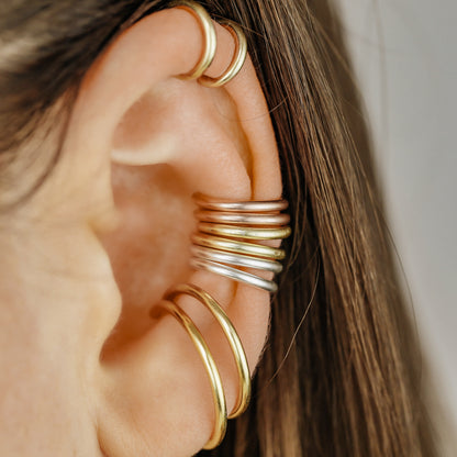 Thick smooth no-piercing ear cuff shown worn on different areas of the ear, including lobe, conch, cartilage and upper helix, with a rounded wrap design available in sterling silver, 18k yellow gold plated sterling silver, 18k rose gold plated sterling silver and black aluminum.