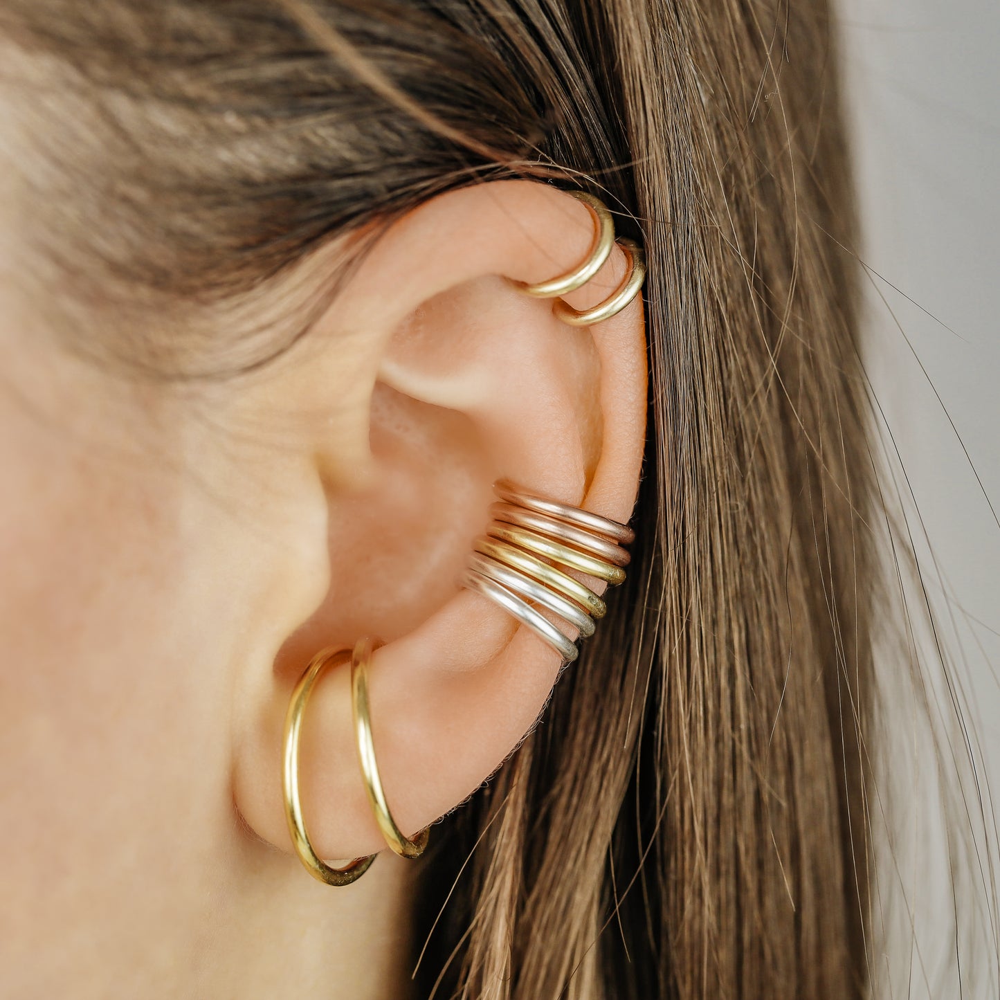 Thick smooth no-piercing ear cuff shown worn on different areas of the ear, including lobe, conch, cartilage and upper helix, with a rounded wrap design available in sterling silver, 18k yellow gold plated sterling silver, 18k rose gold plated sterling silver and black aluminum.