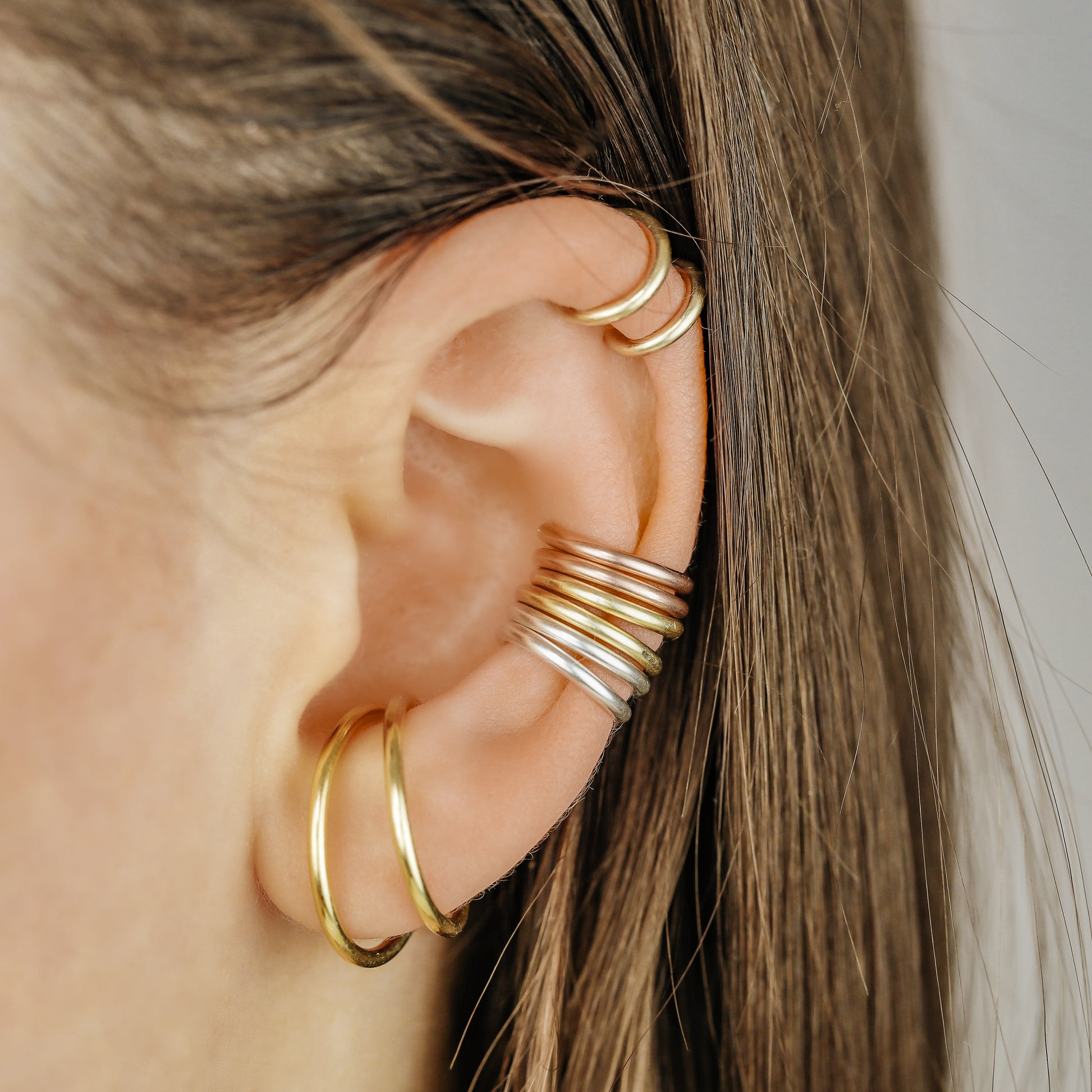 Thick smooth no-piercing ear cuff shown worn on different areas of the ear, including lobe, conch, cartilage and upper helix, with a rounded wrap design available in sterling silver, 18k yellow gold plated sterling silver, 18k rose gold plated sterling silver and black aluminum.