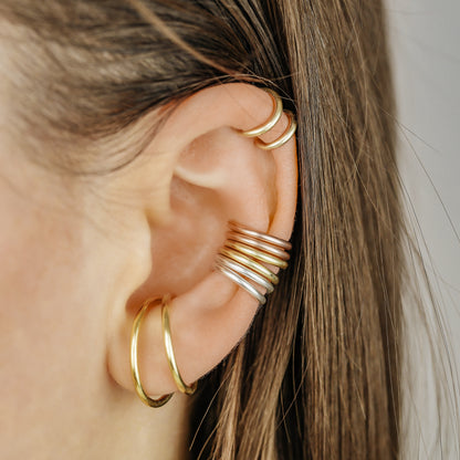 Thick smooth no-piercing ear cuff shown worn on different areas of the ear, including lobe, conch, cartilage and upper helix, with a rounded wrap design available in sterling silver, 18k yellow gold plated sterling silver, 18k rose gold plated sterling silver and black aluminum.