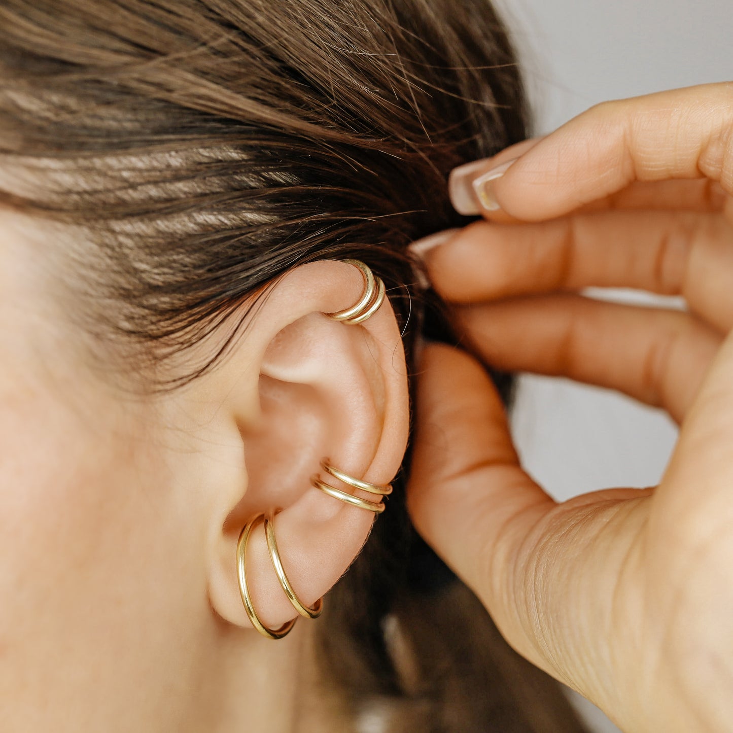 Thick smooth no-piercing ear cuff shown worn on different areas of the ear, including lobe, conch, cartilage and upper helix, with a rounded wrap design available in sterling silver, 18k yellow gold plated sterling silver, 18k rose gold plated sterling silver and black aluminum.