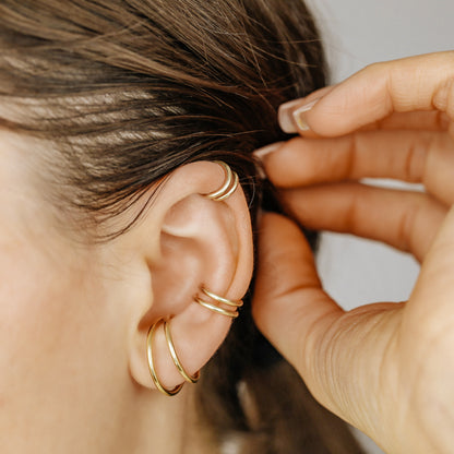 Thick smooth no-piercing ear cuff shown worn on different areas of the ear, including lobe, conch, cartilage and upper helix, with a rounded wrap design available in sterling silver, 18k yellow gold plated sterling silver, 18k rose gold plated sterling silver and black aluminum.