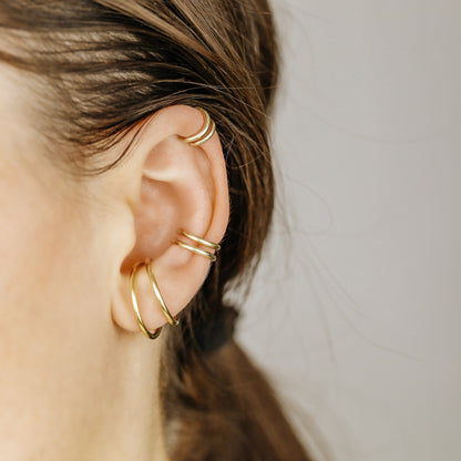 Thick smooth no-piercing ear cuff shown worn on different areas of the ear, including lobe, conch, cartilage and upper helix, with a rounded wrap design available in sterling silver, 18k yellow gold plated sterling silver, 18k rose gold plated sterling silver and black aluminum.