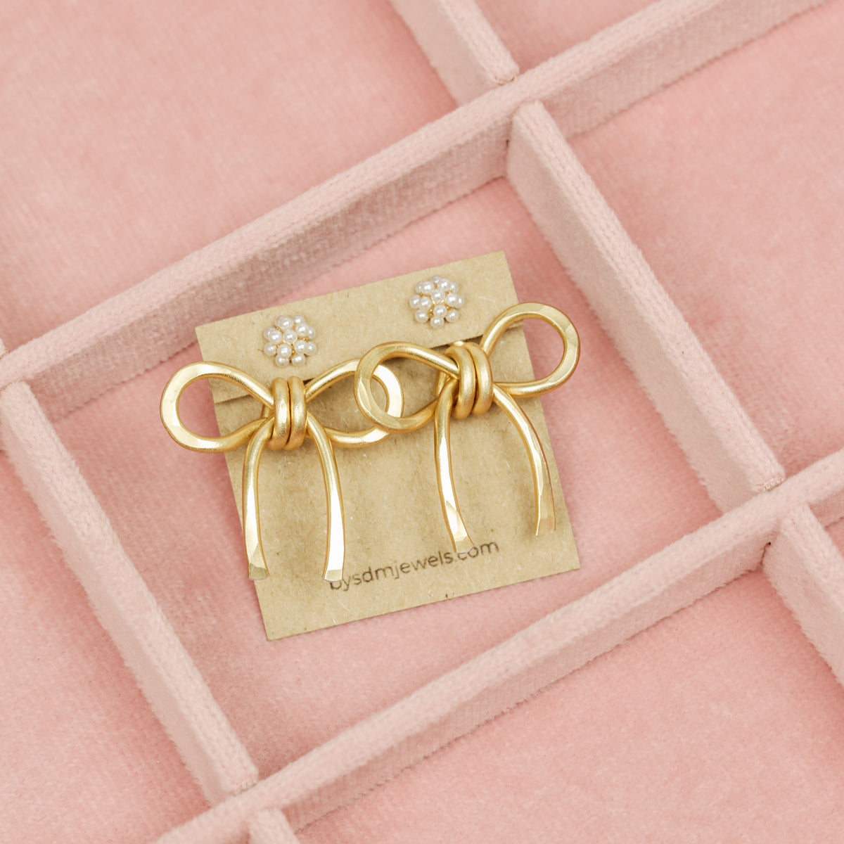 Pair of gold medium bow ear jackets displayed on a card inside a pink tray, styled with pearl cluster stud earrings.