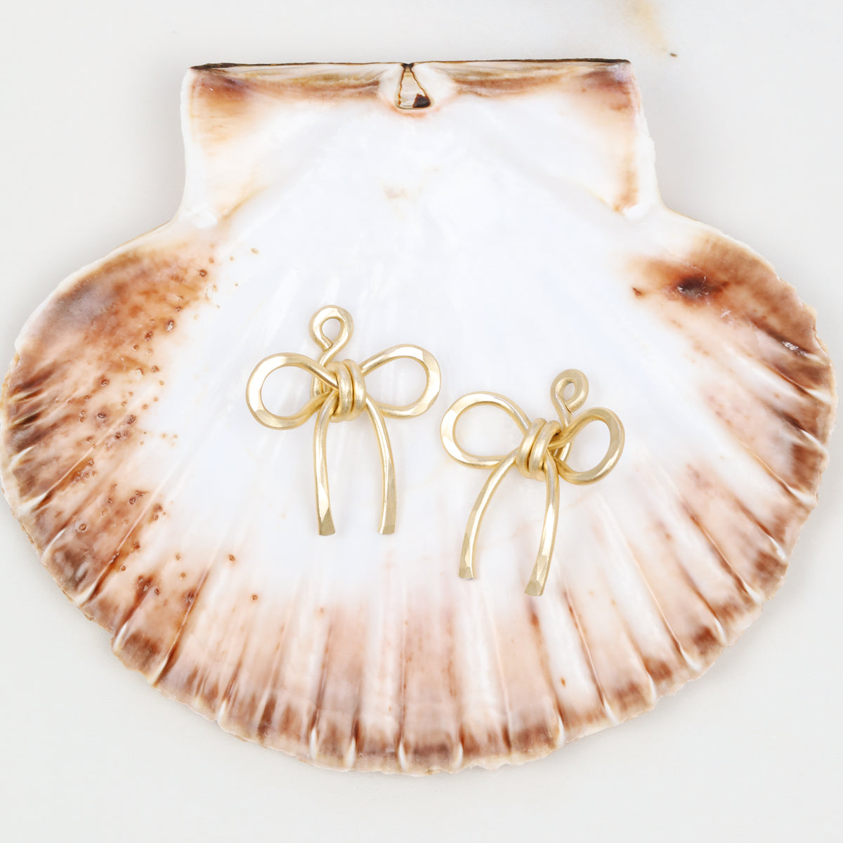 Pair of medium hammered aluminum bow ear jackets in gold displayed on a large seashell, showing the sculptural bow shape and medium-size silhouette.
