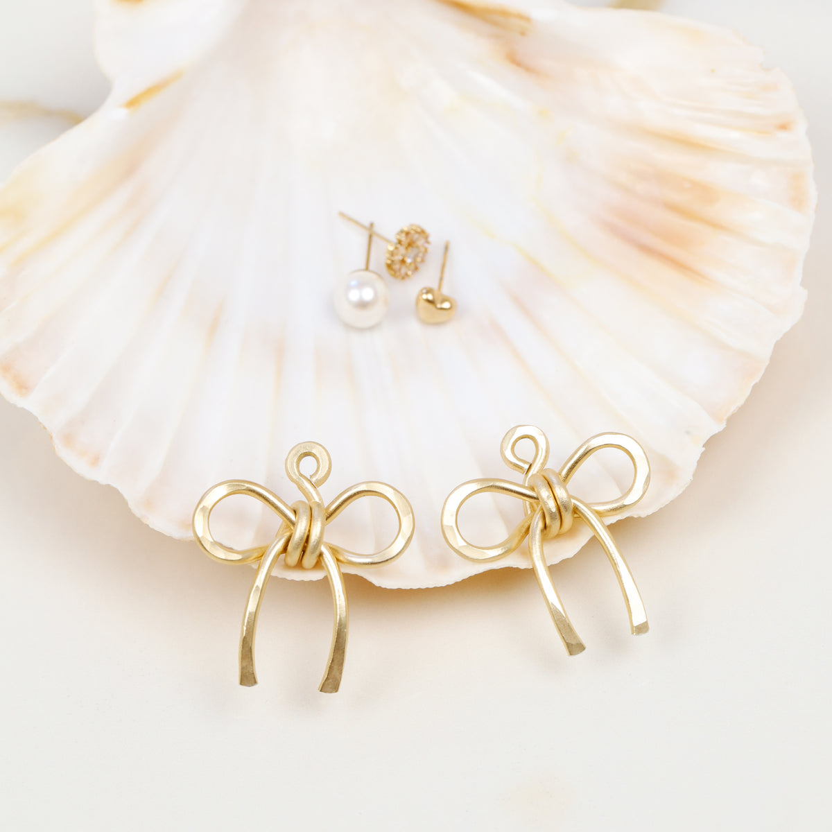 Gold medium bow ear jackets photographed in front of a seashell with example stud earrings in the background, showing the interchangeable ear jacket concept.