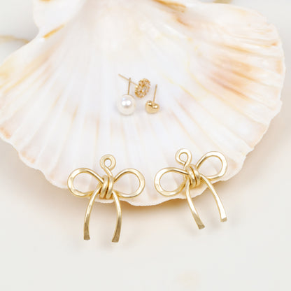 Gold medium bow ear jackets photographed in front of a seashell with example stud earrings in the background, showing the interchangeable ear jacket concept.
