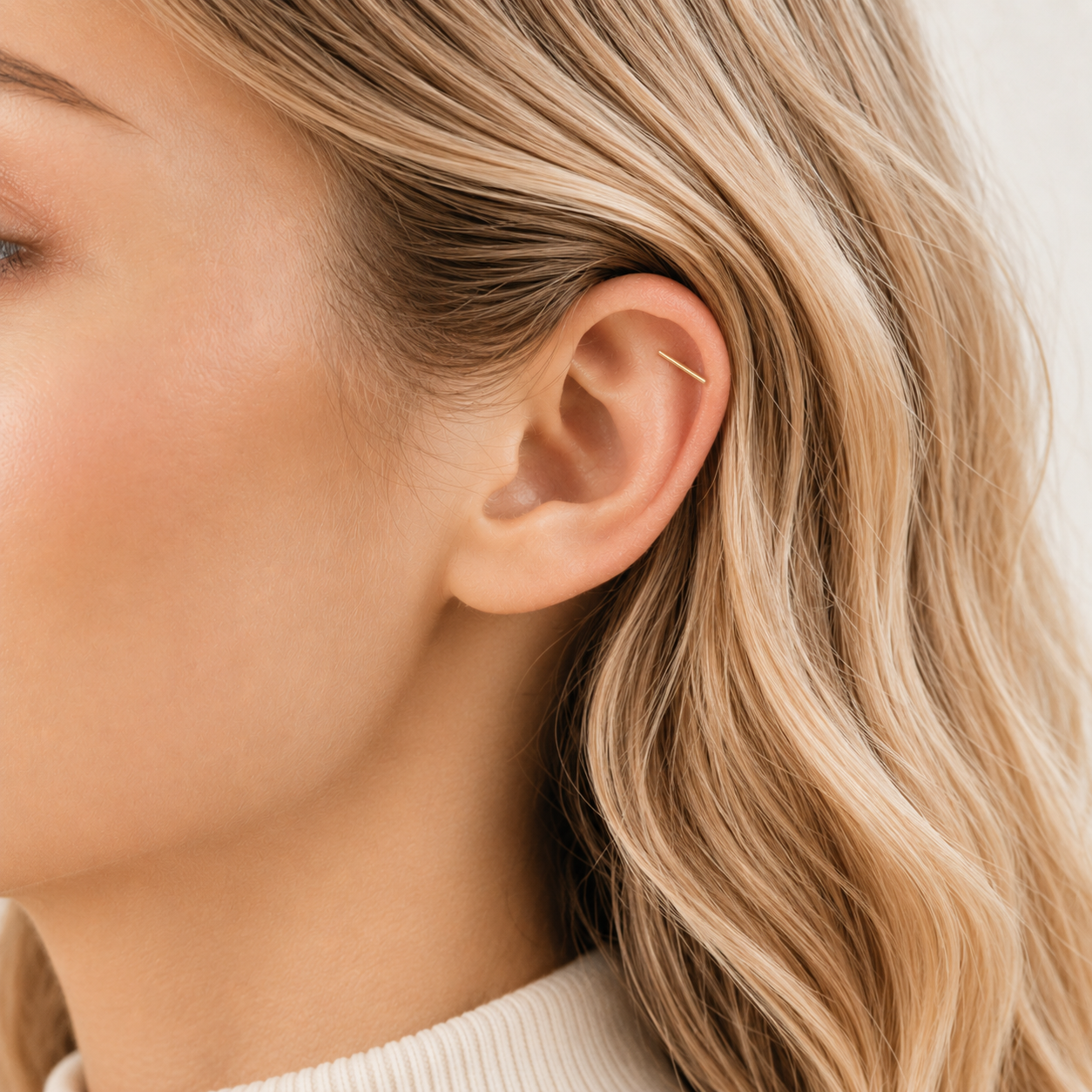 Blonde woman with soft wavy hair wearing a minimal gold bar helix earring on upper cartilage piercing close up side profile