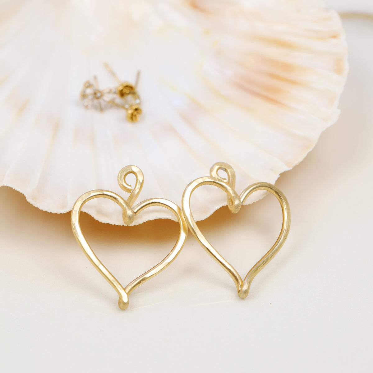 Close-up of a pair of handmade gold Large Heart Ear Jackets displayed in front of a natural shell, showing the open elongated heart shape, hammered surface and curved top detail.