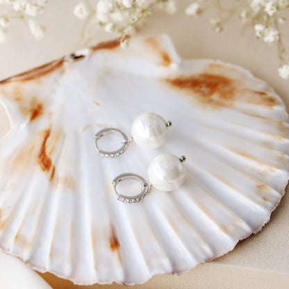 Pearl drop hoop earrings displayed on a shell with round ceramic pearls and sparkling hoops