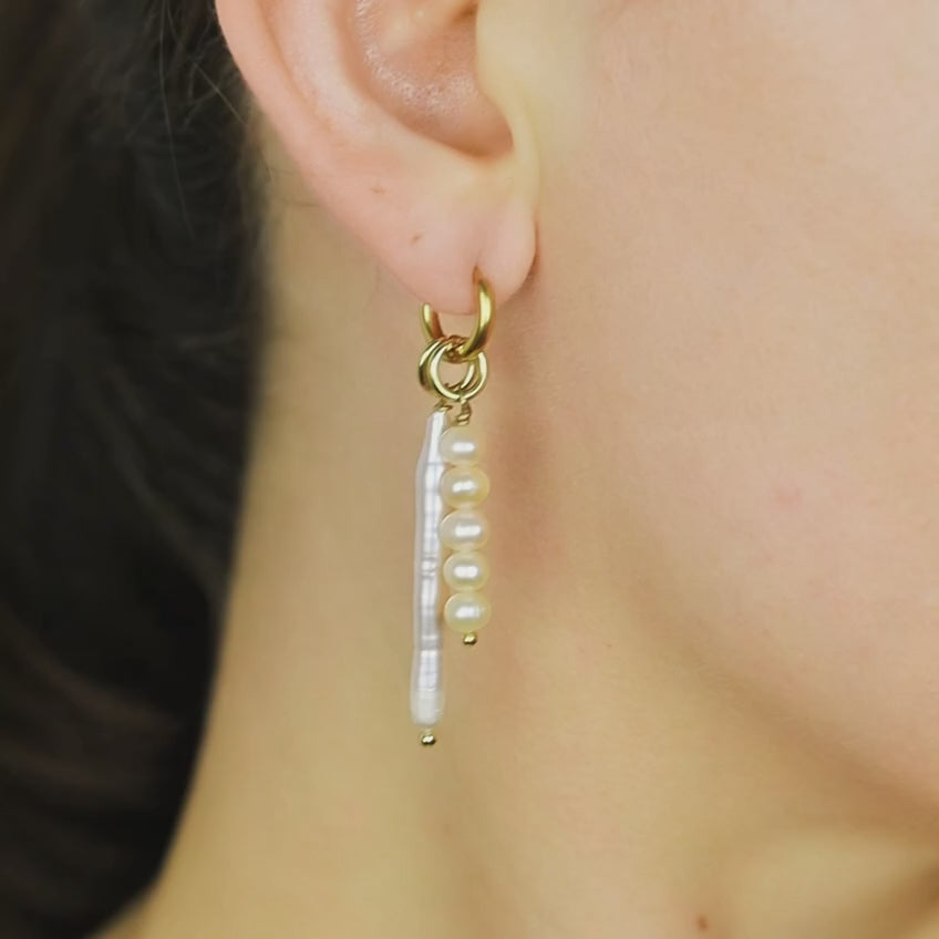 Close-up of a woman wearing an 18k gold plated huggie hoop earring adorned with a long pink baroque stick pearl and a matching five-pearl cluster charm.