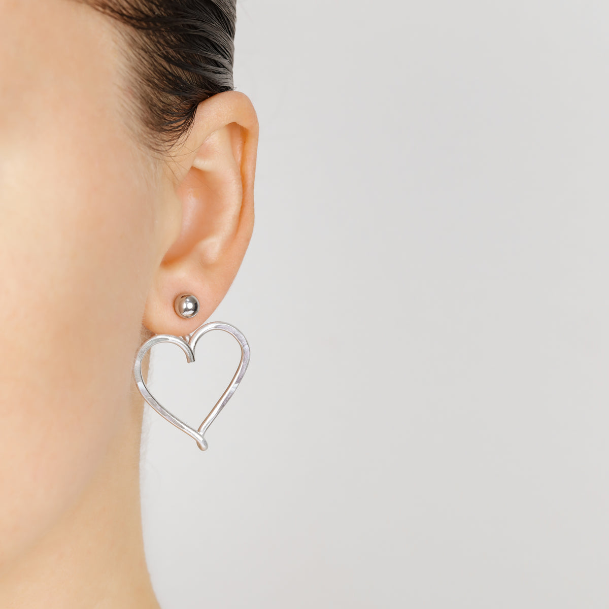Silver Large Heart Ear Jacket worn behind the earlobe with a polished ball stud, showing the elongated heart outline and two-part Ear Jacket construction.
