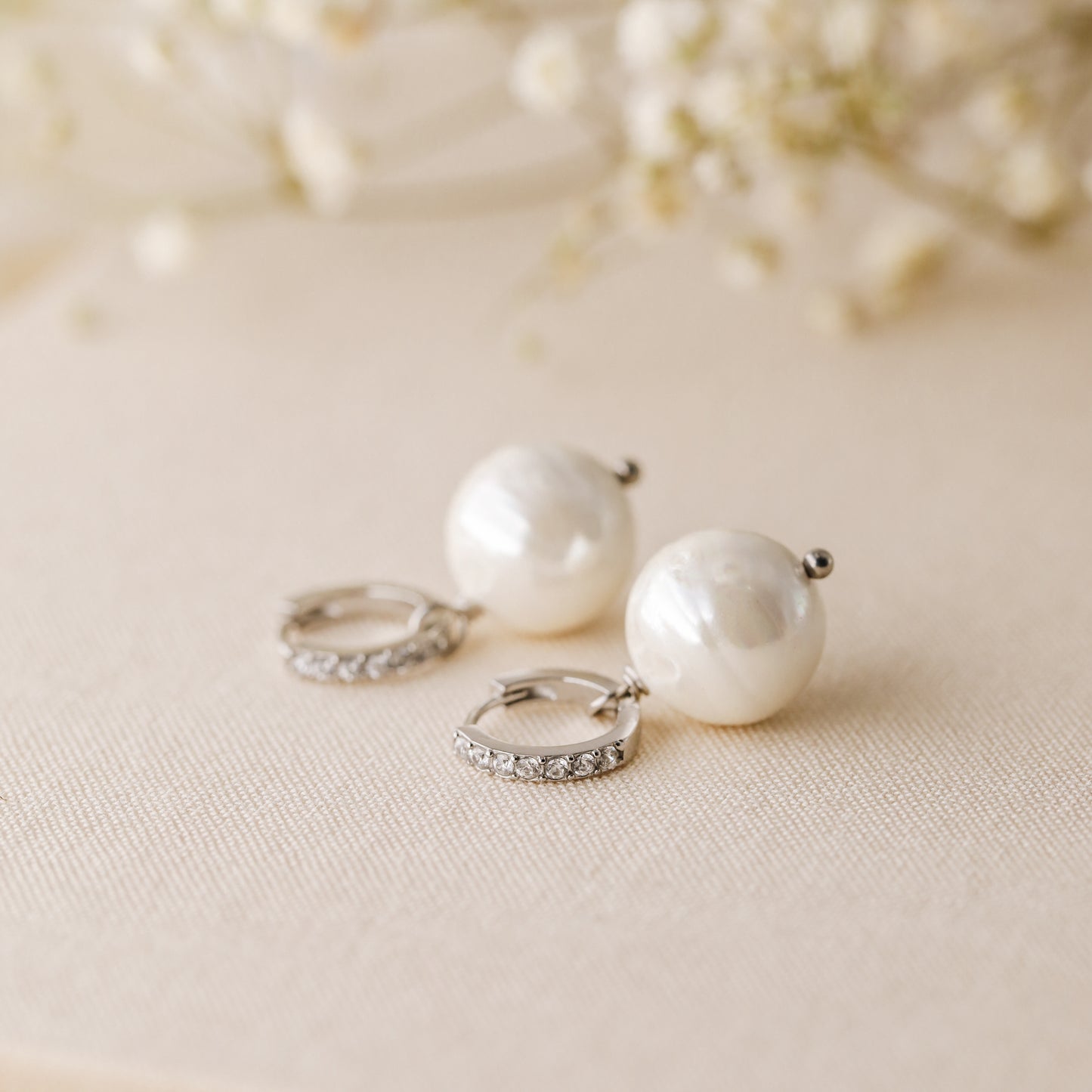Silver pearl drop hoop earrings styled on neutral fabric with soft floral background