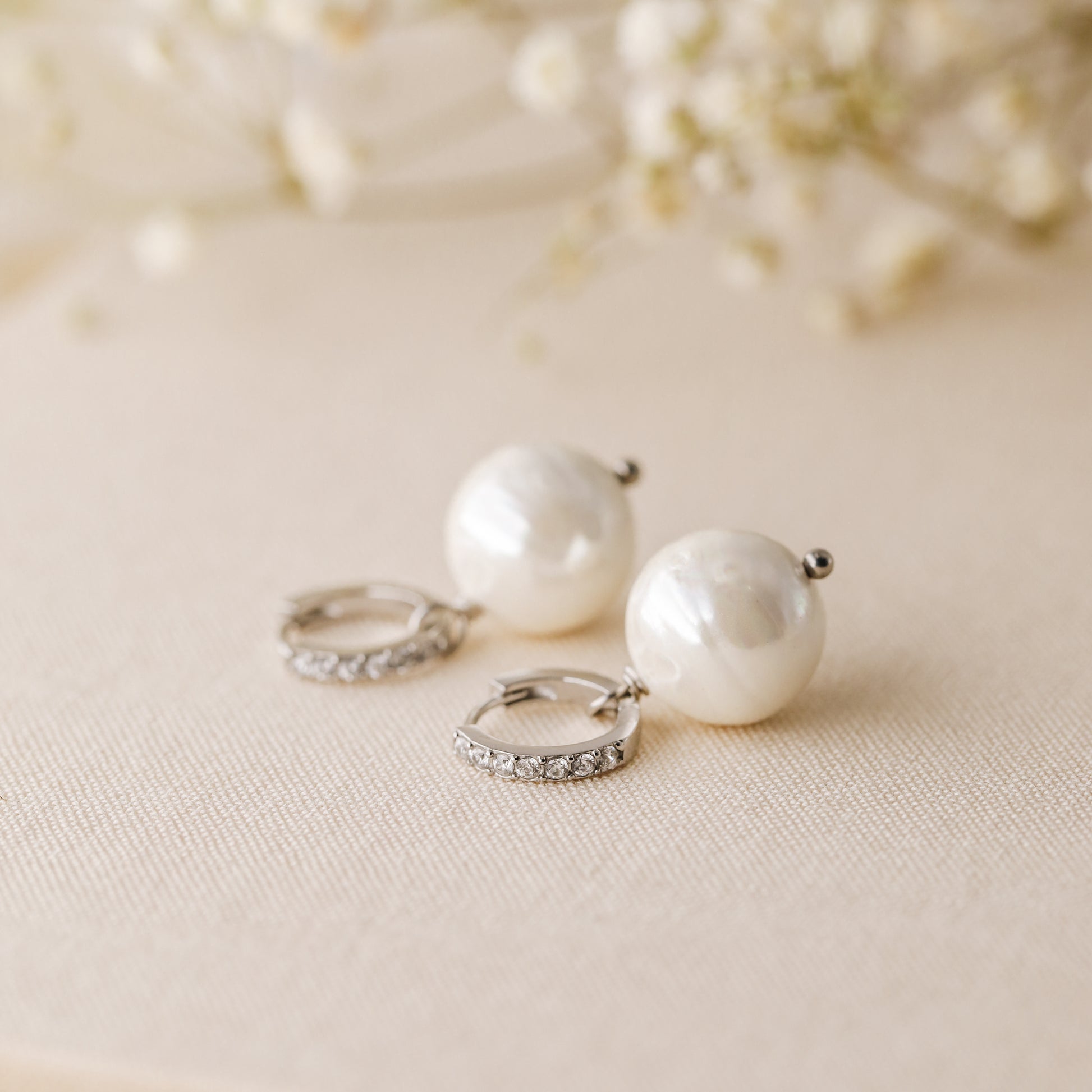 Silver pearl drop hoop earrings styled on neutral fabric with soft floral background