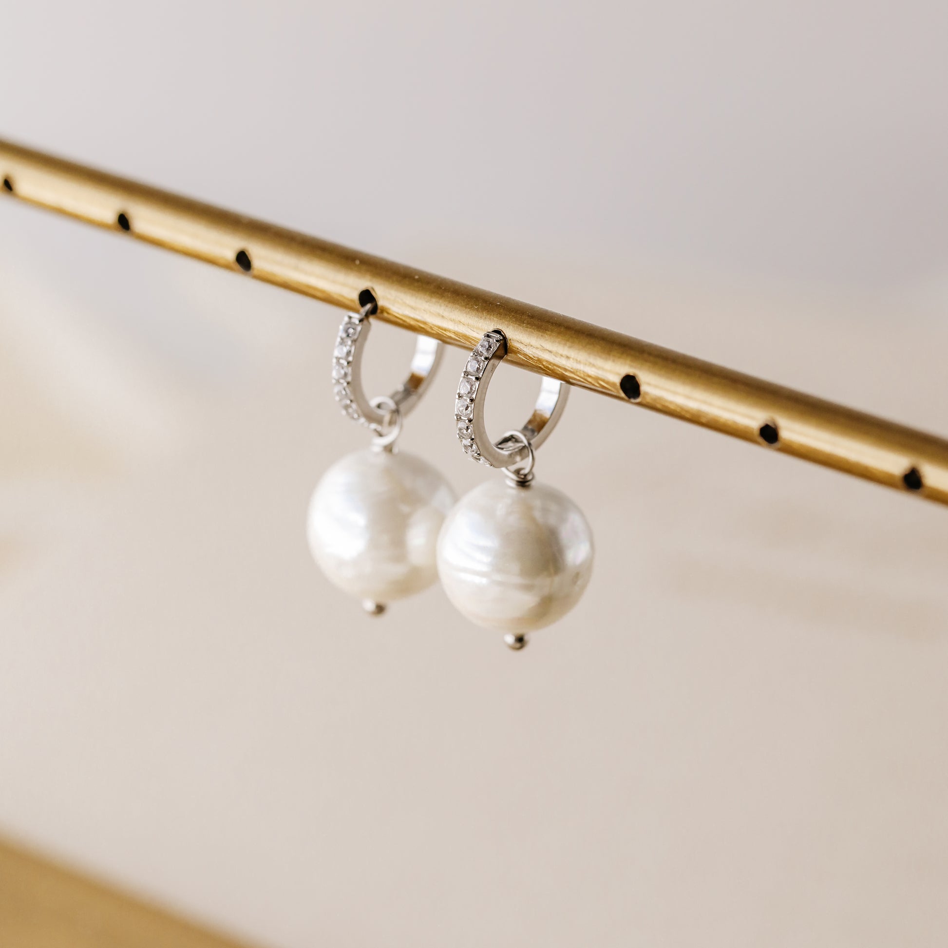 Silver pearl drop hoop earrings hanging from a gold display bar with sparkling hoop detail