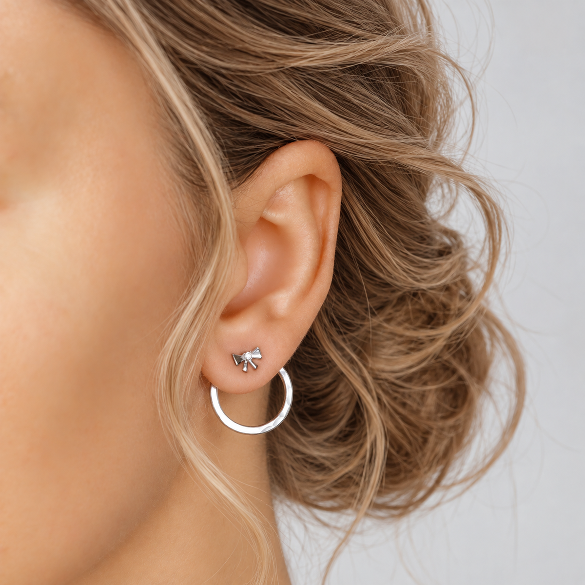 Silver round ear jacket worn behind the lobe with a small bow stud, showing the circular hammered jacket hanging below the ear.