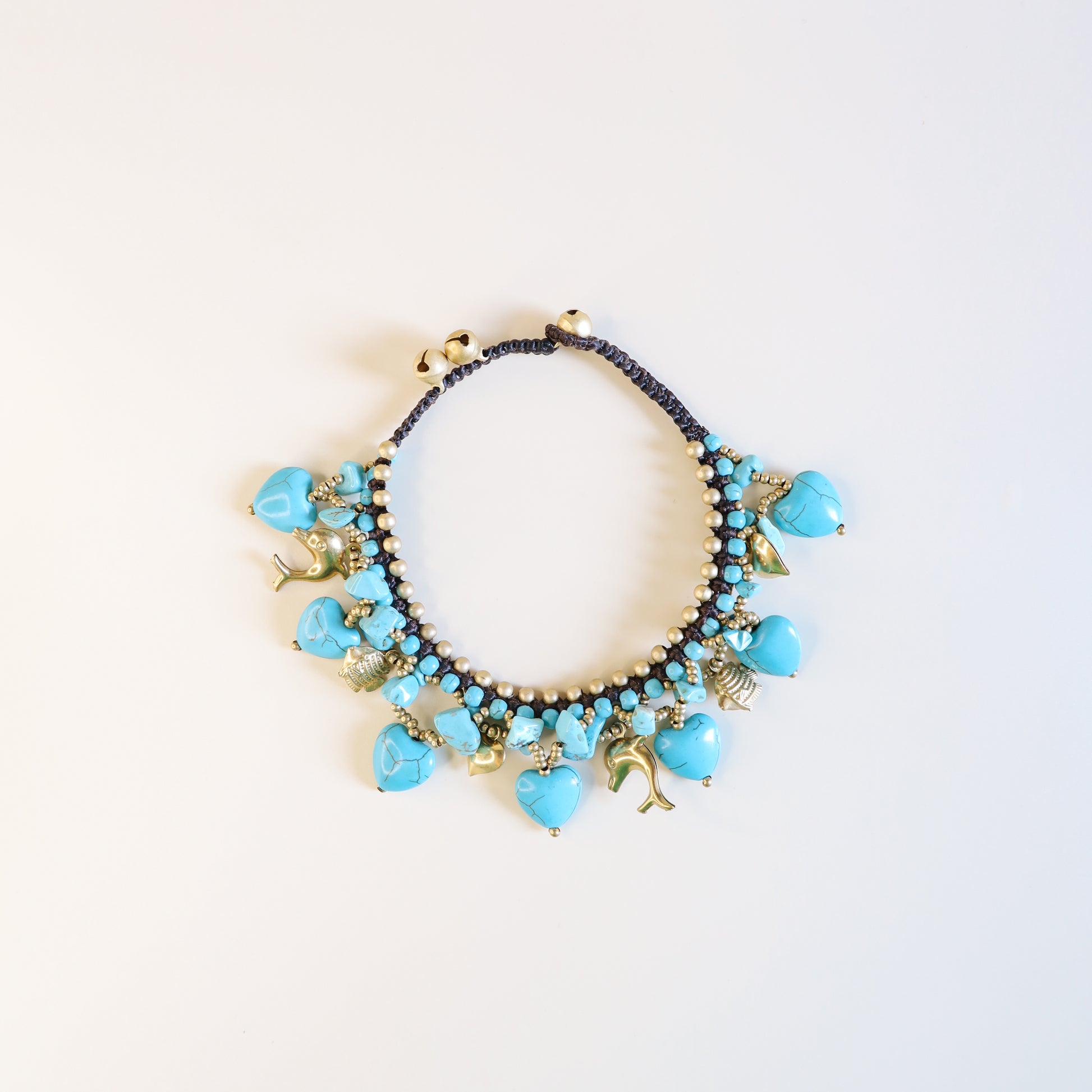 Close-up of adjustable turquoise anklet with brown cord, brass beads, heart charms, dolphins, fish and lucky bells.