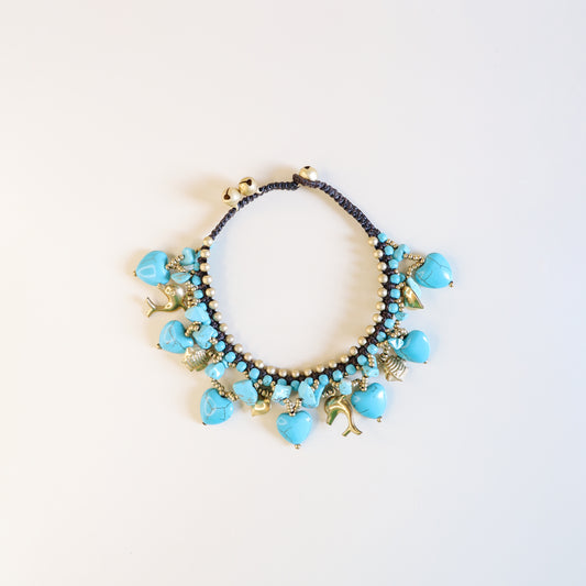 Close-up of adjustable turquoise anklet with brown cord, brass beads, heart charms, dolphins, fish and lucky bells.